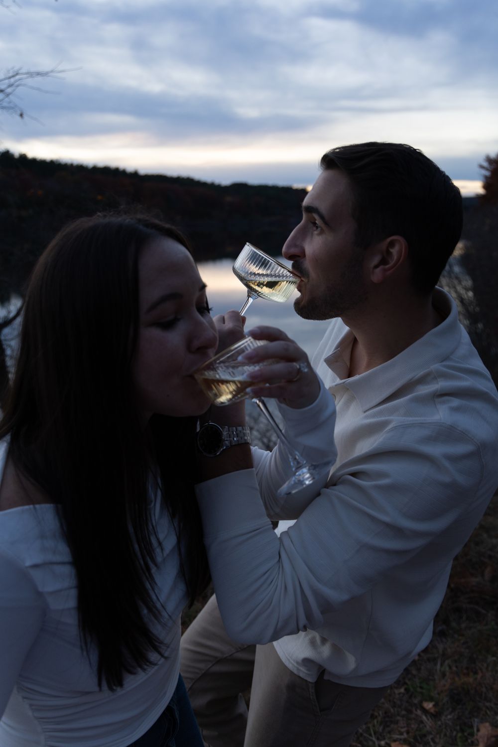 Romantic couple drinking champagne together by a lake at dusk in white outfits.