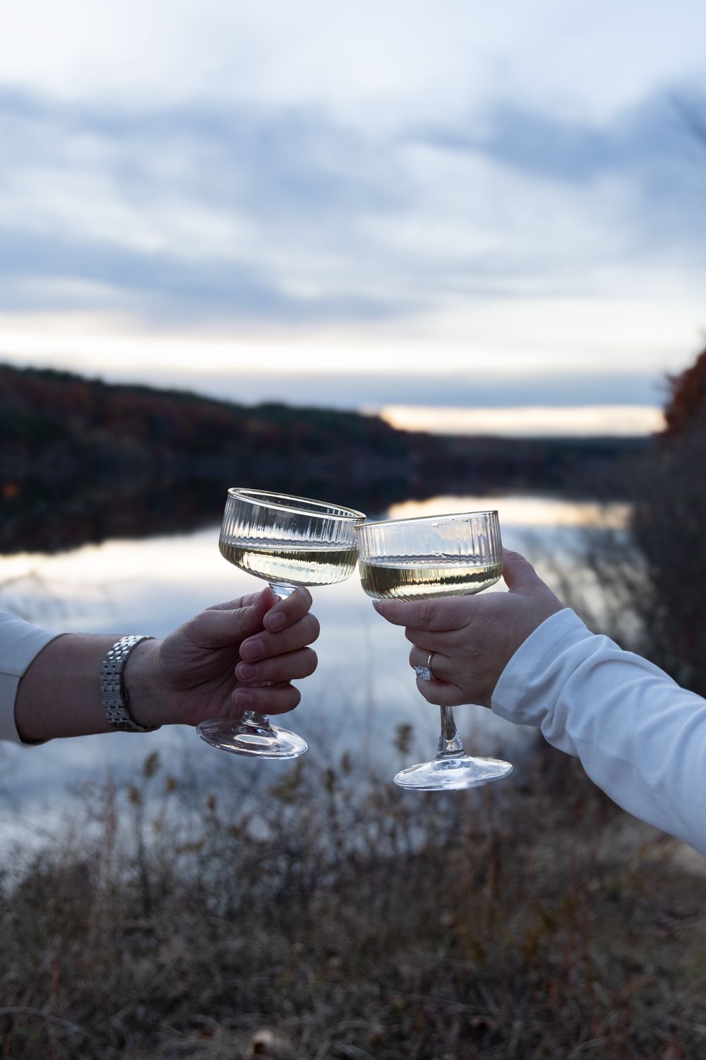 Couple clinking champagne coupe glasses outdoors by a reflective lake at dusk.
