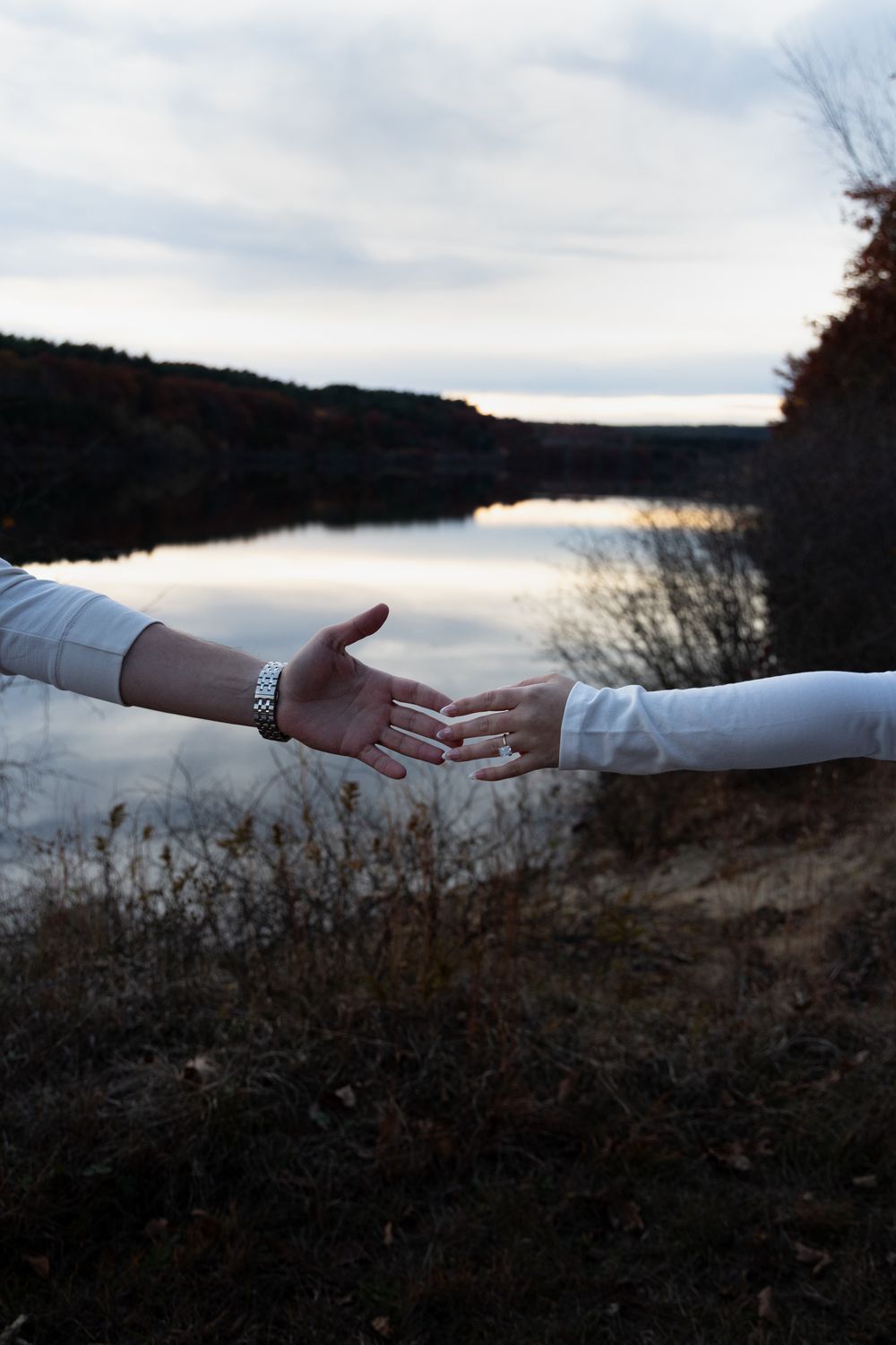 Two people reaching out to hold hands near a tranquil lake at twilight in white shirts.