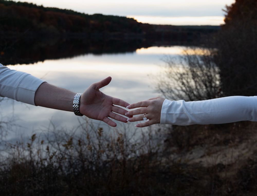Couple nearly touching hands near a calm lake at dusk, both wearing white long-sleeve shirts.