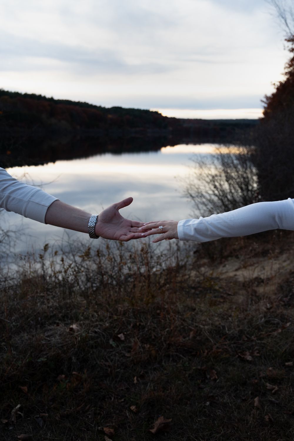 Couple extending hands to each other by a serene lake at dusk, dressed in white shirts.