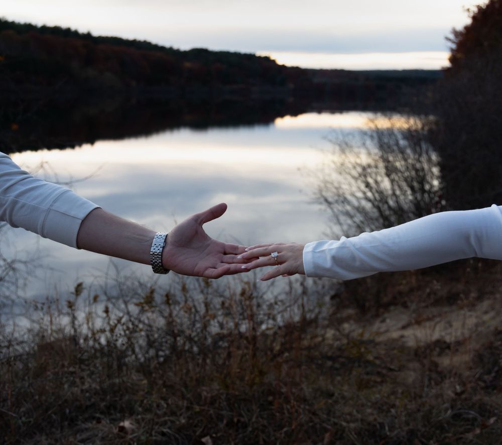 Two people reaching toward each other near a calm lake at twilight, wearing white shirts.