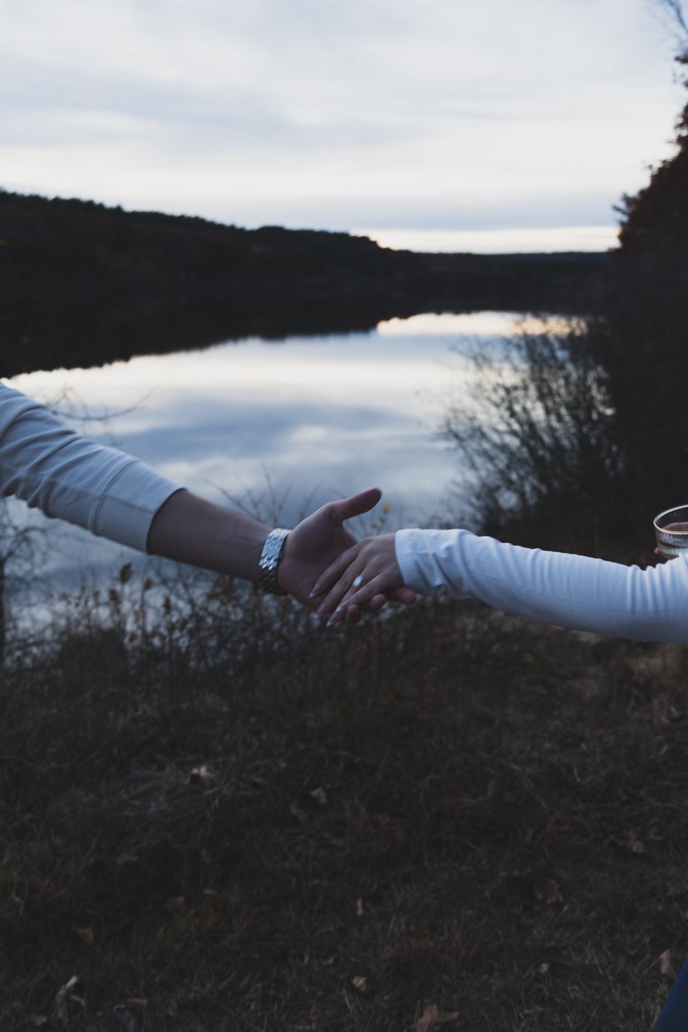 Couple holding hands near a reflective lake at dusk, both wearing white long-sleeve shirts.
