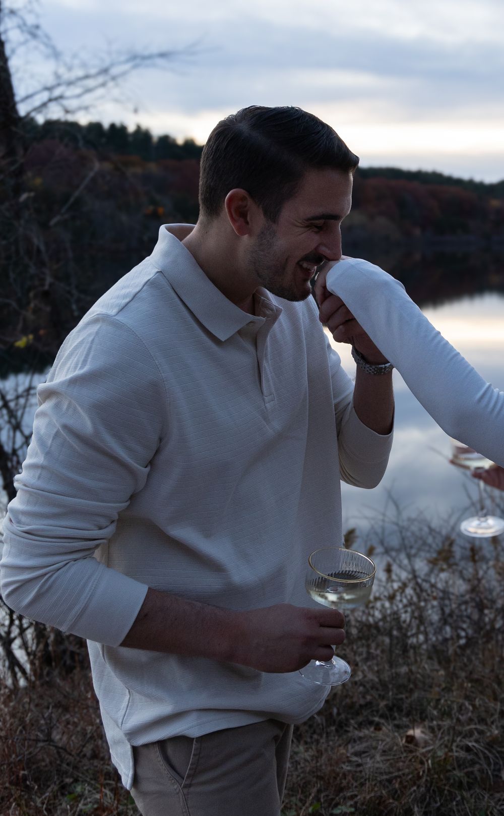 Smiling man in white polo shirt holding champagne glass by a lake at sunset.