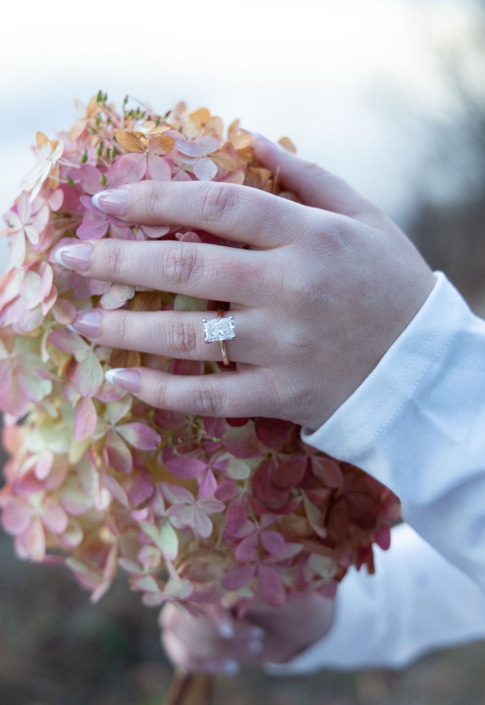 Close-up of an engagement ring on a hand holding pink hydrangea flowers outdoors.