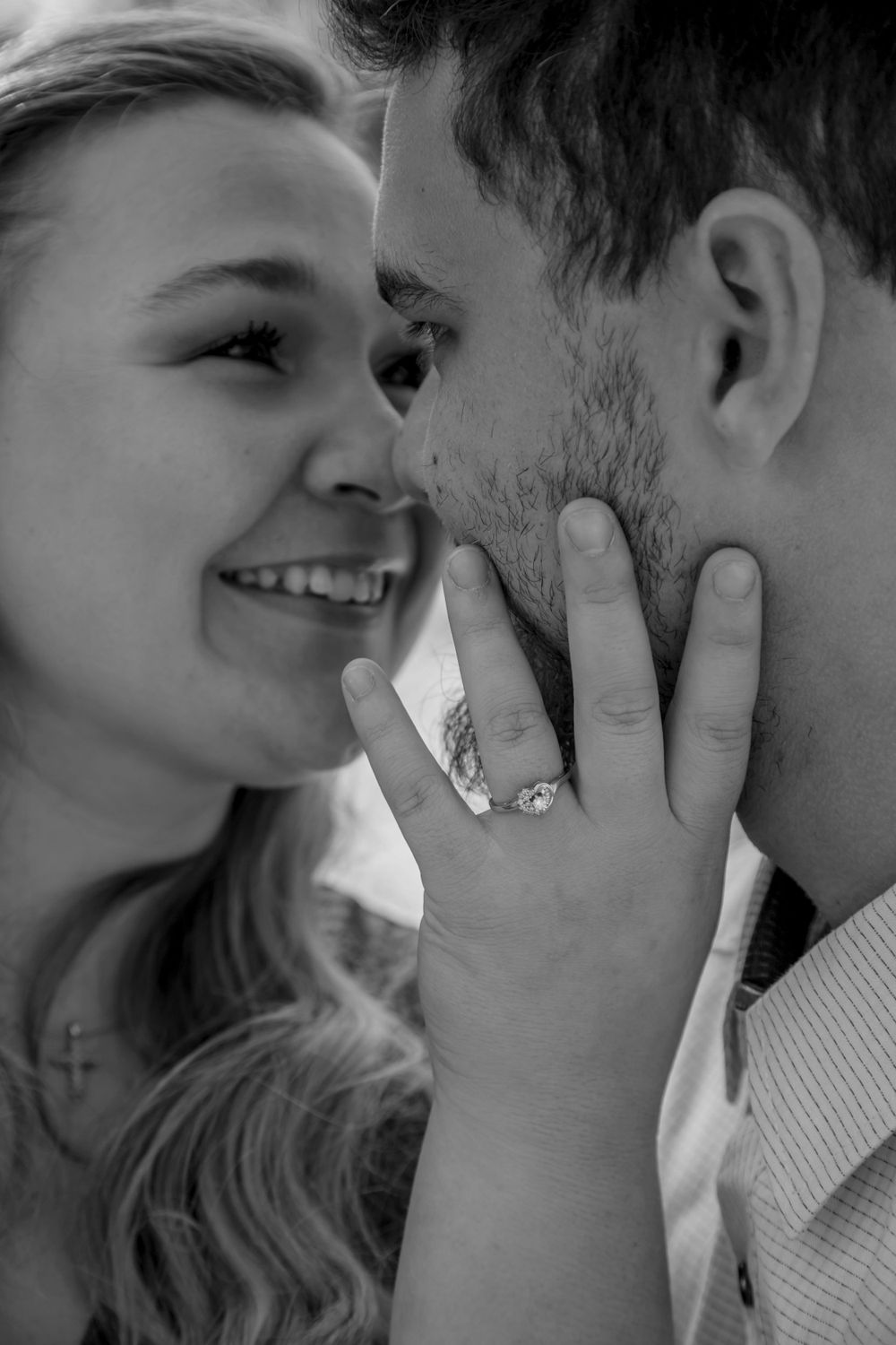 Black and white close-up of smiling woman showing engagement ring while touching mans face.
