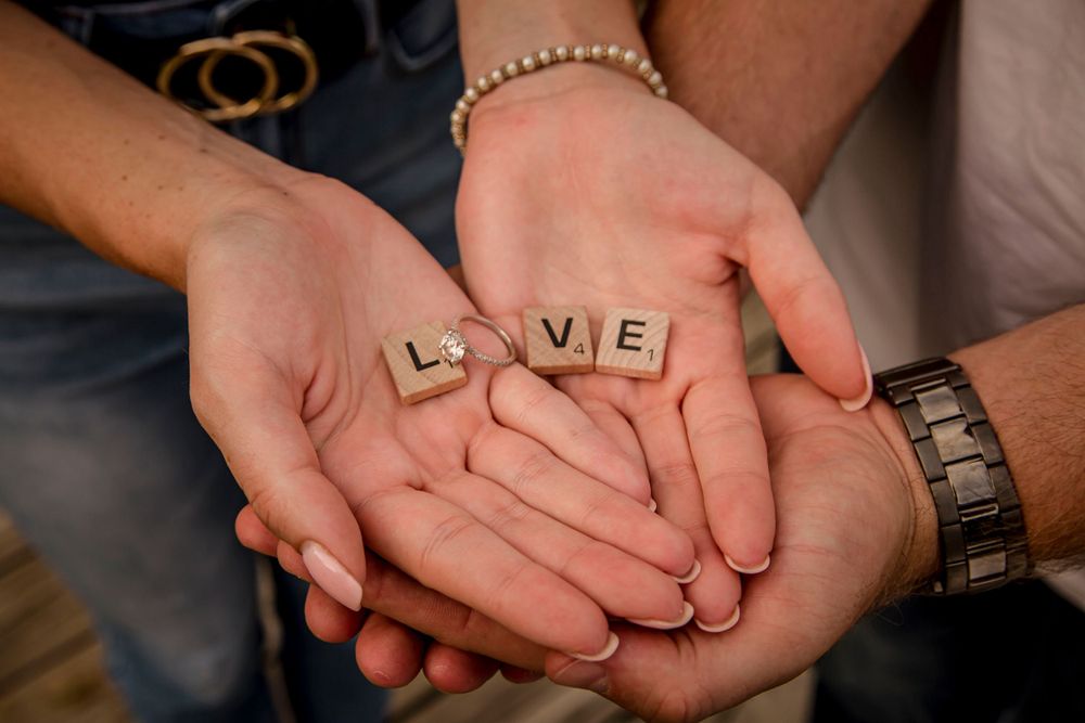 Two people holding Scrabble tiles spelling LOVE in their cupped hands, wearing bracelets and a watch.