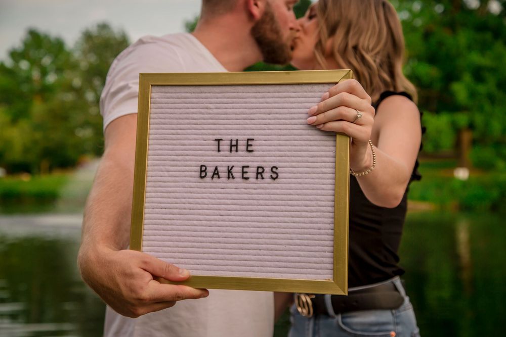 Engaged couple kissing behind a letter board sign reading The Bakers near a scenic outdoor pond.