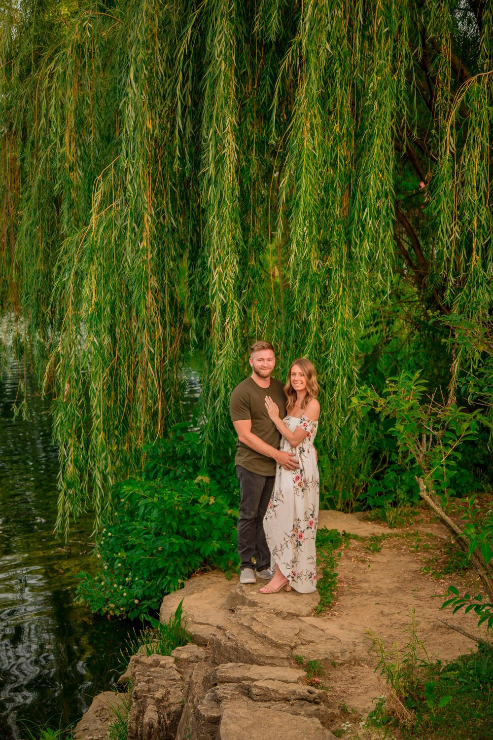 Couple embracing romantically under a weeping willow tree beside a tranquil pond in a lush green park.