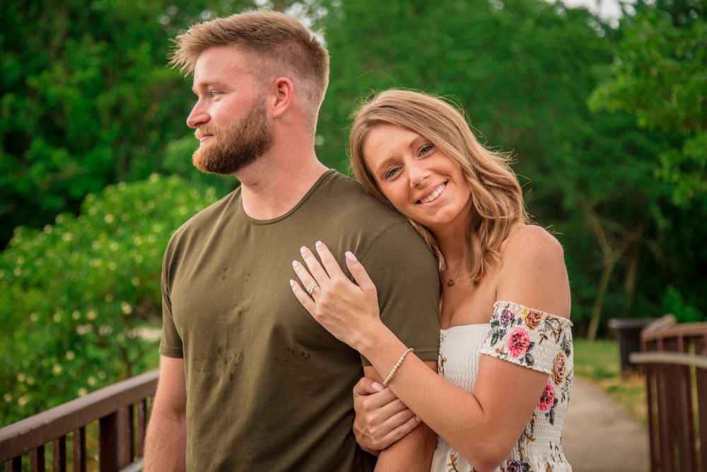 Smiling couple posing on a wooden bridge outdoors, woman hugging man from behind in a lush green park.
