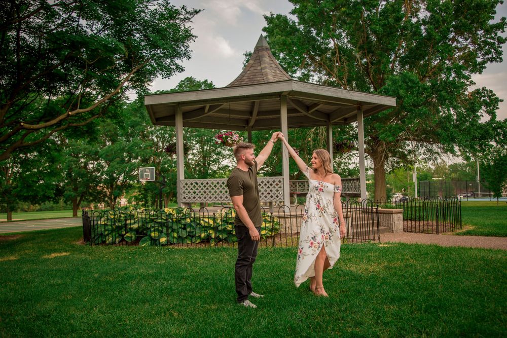 Joyful couple dancing and spinning near a charming gazebo surrounded by lush green trees in a park.