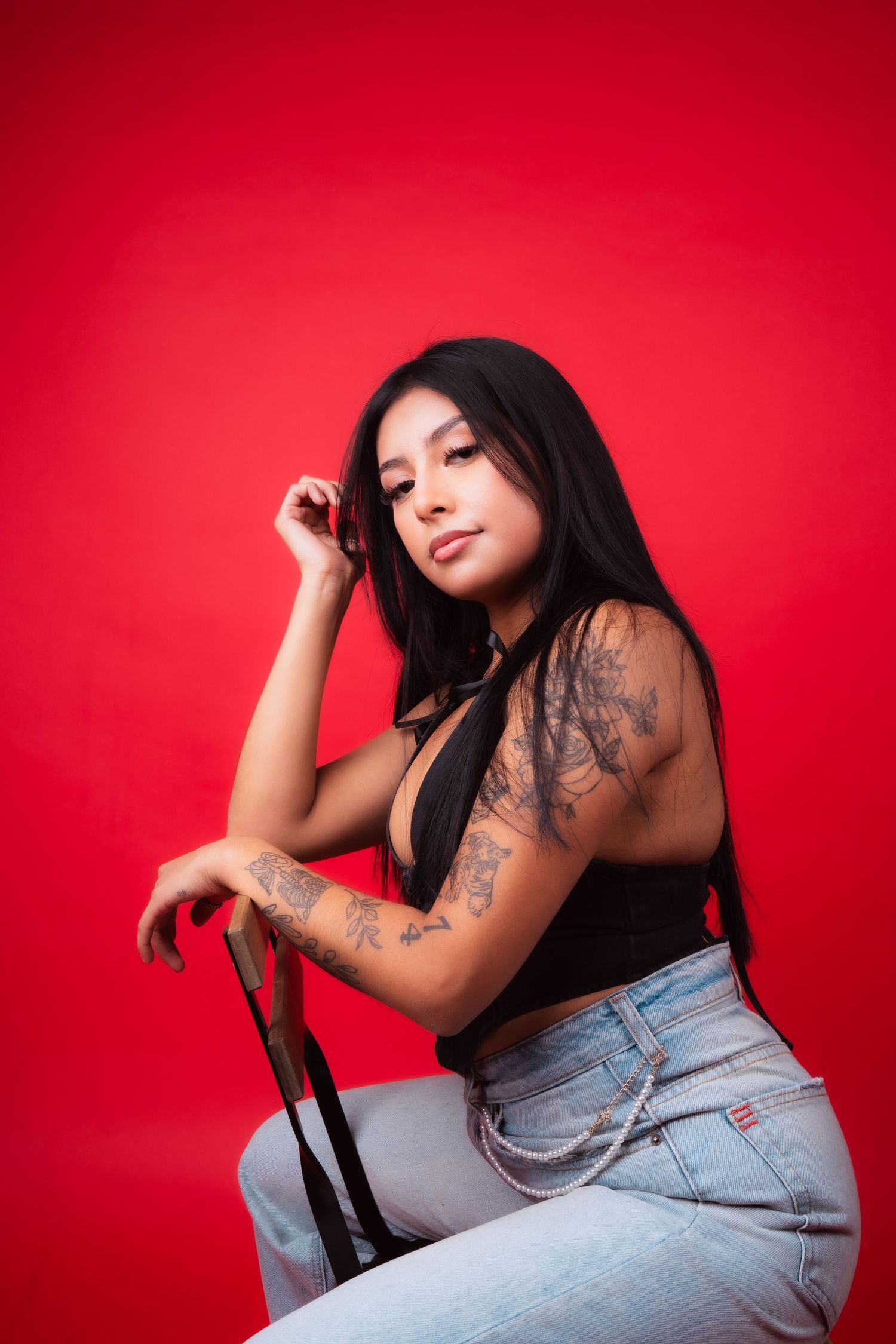 Model in black top and jeans poses against vibrant red background showing tattoo art.