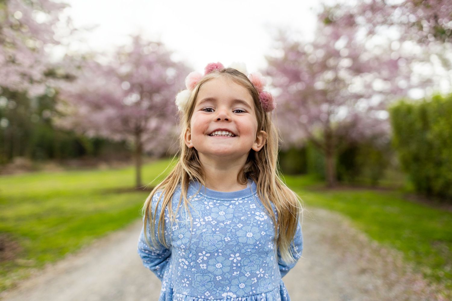A happy child wearing a blue floral dress stands on a gravel path with blooming cherry trees in the background.