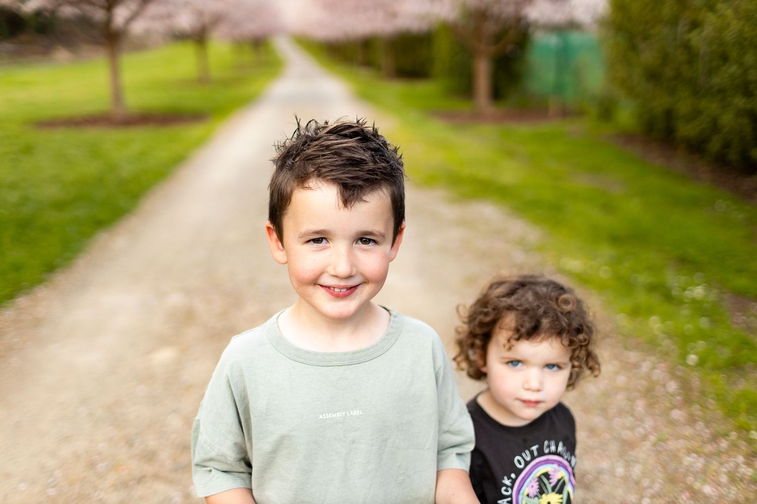 Two young children stand together on a gravel path with green grass and trees in the background.
