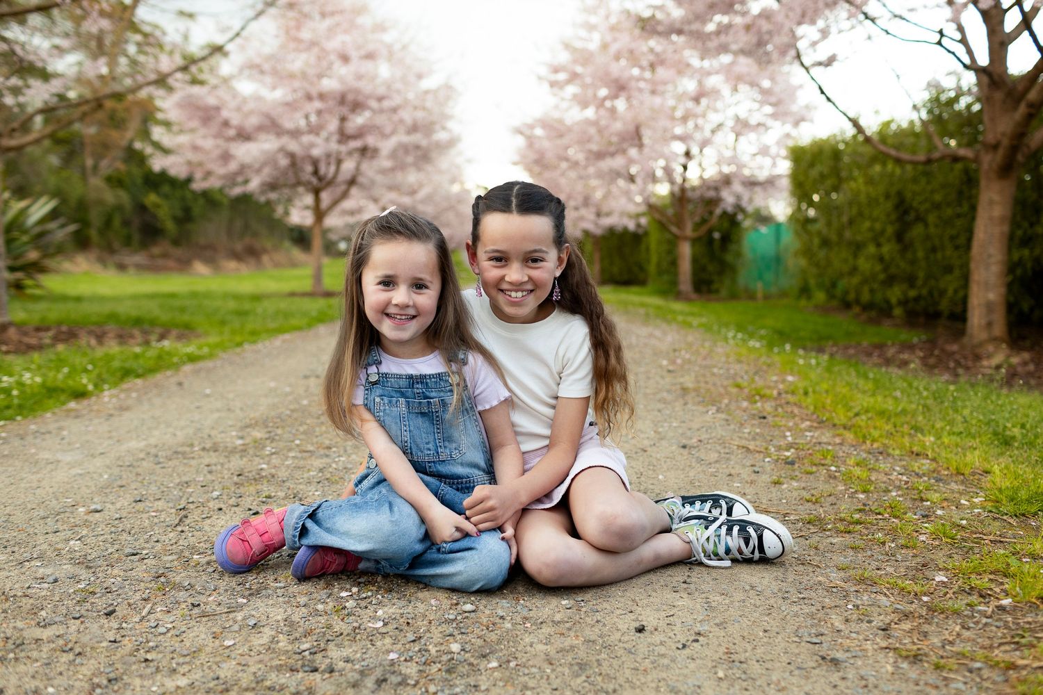 Two young children sitting together on a dirt path with blooming cherry blossom trees in the background.