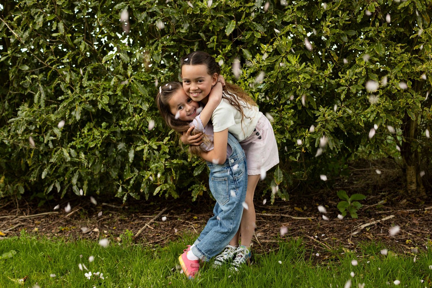 A playful scene in a garden with two sisters hugging each other near blooming trees on a sunny day.