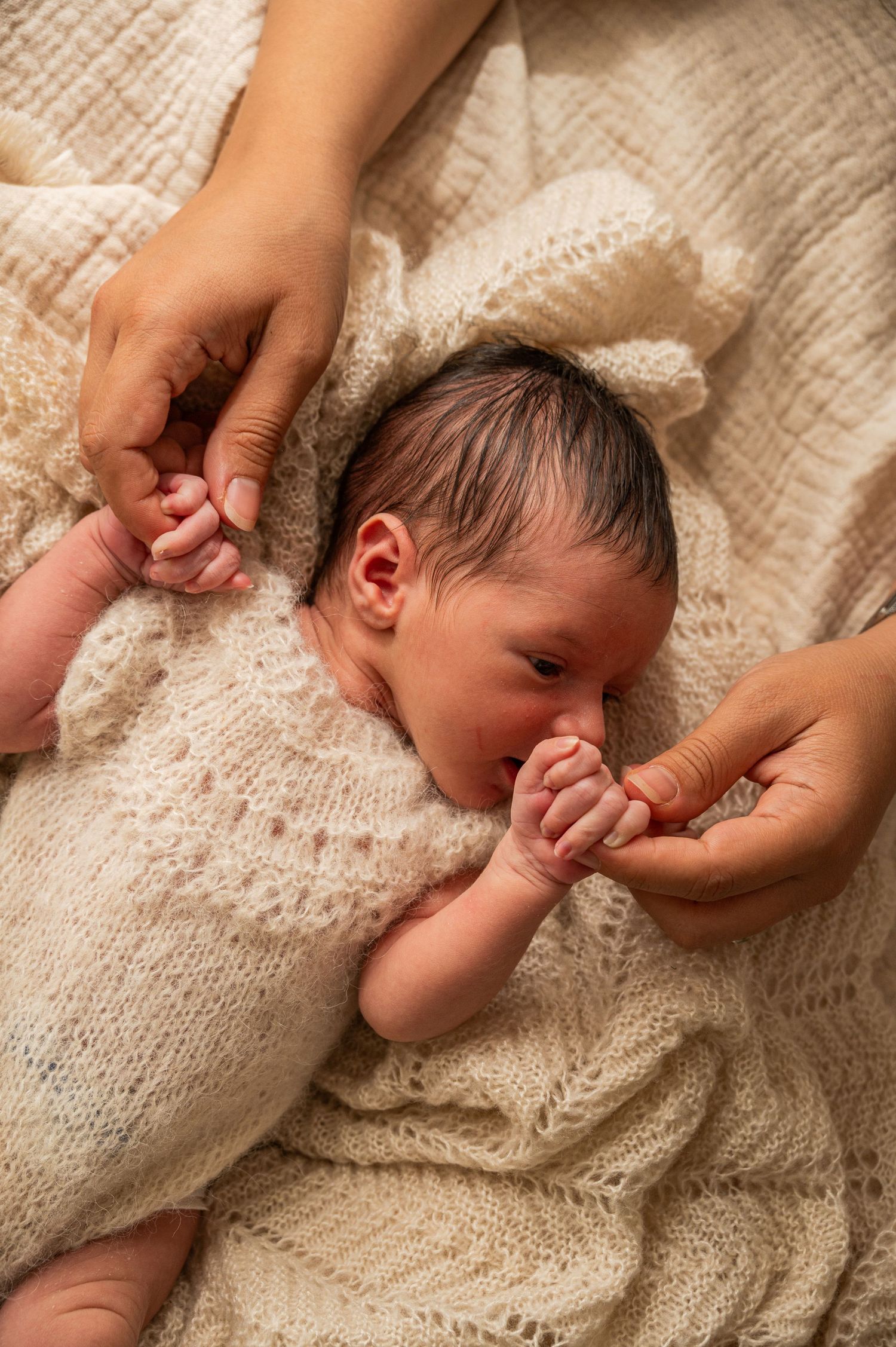 Newborn baby in white knit blanket reaching for parent's hands.