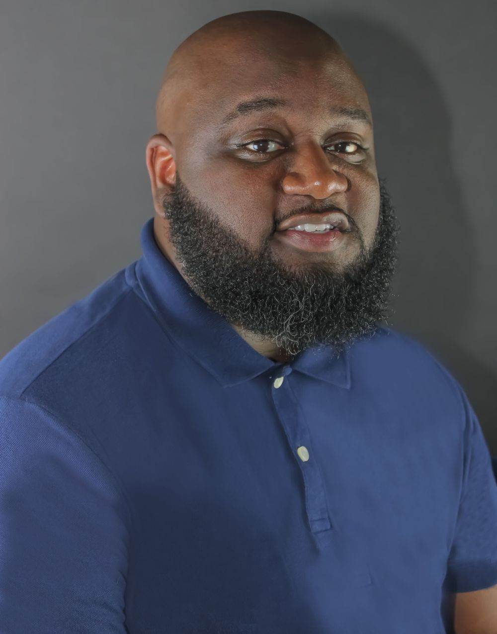 Close-up portrait of person in blue polo shirt with full beard against grey backdrop.