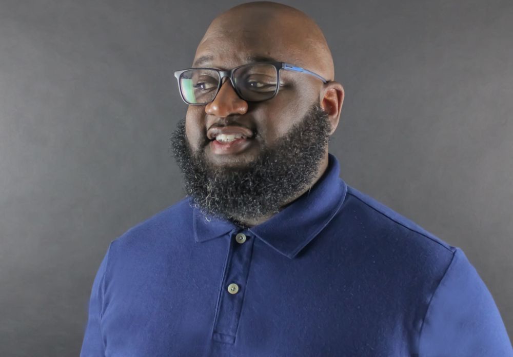 Portrait of person wearing glasses and navy polo shirt with full beard against dark background.