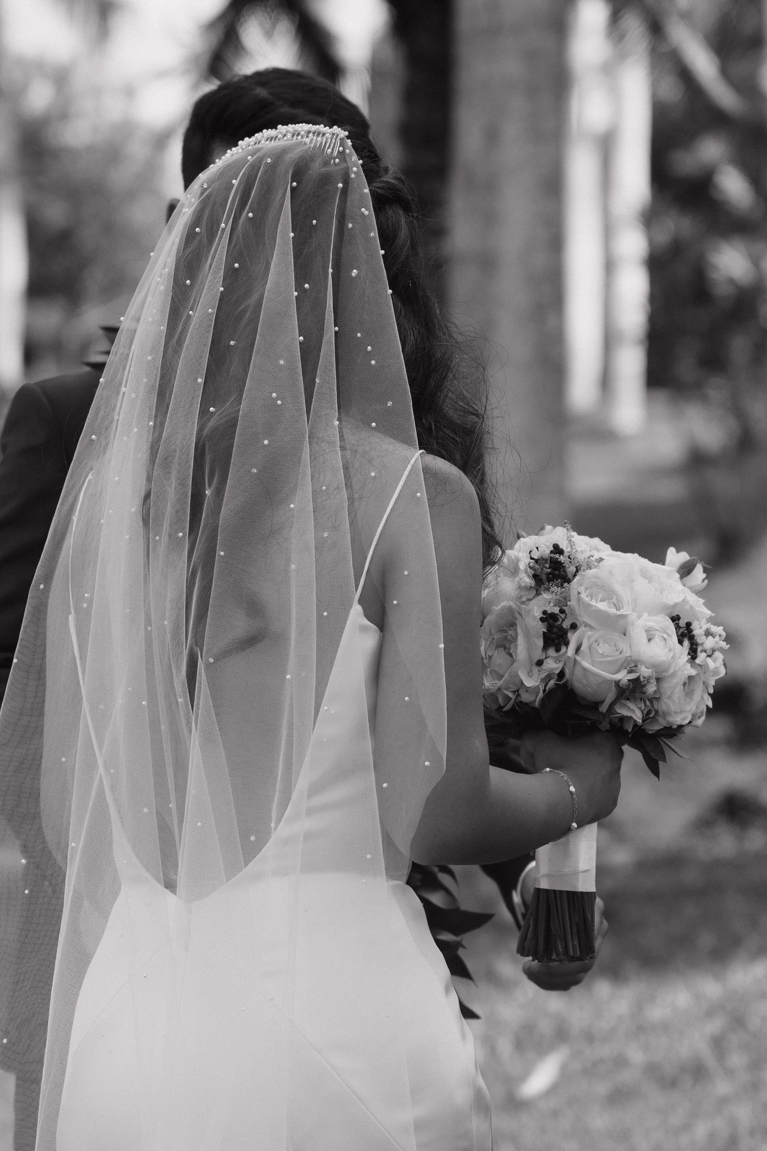 Bride in white dress with pearl-detailed veil holds bouquet from behind.