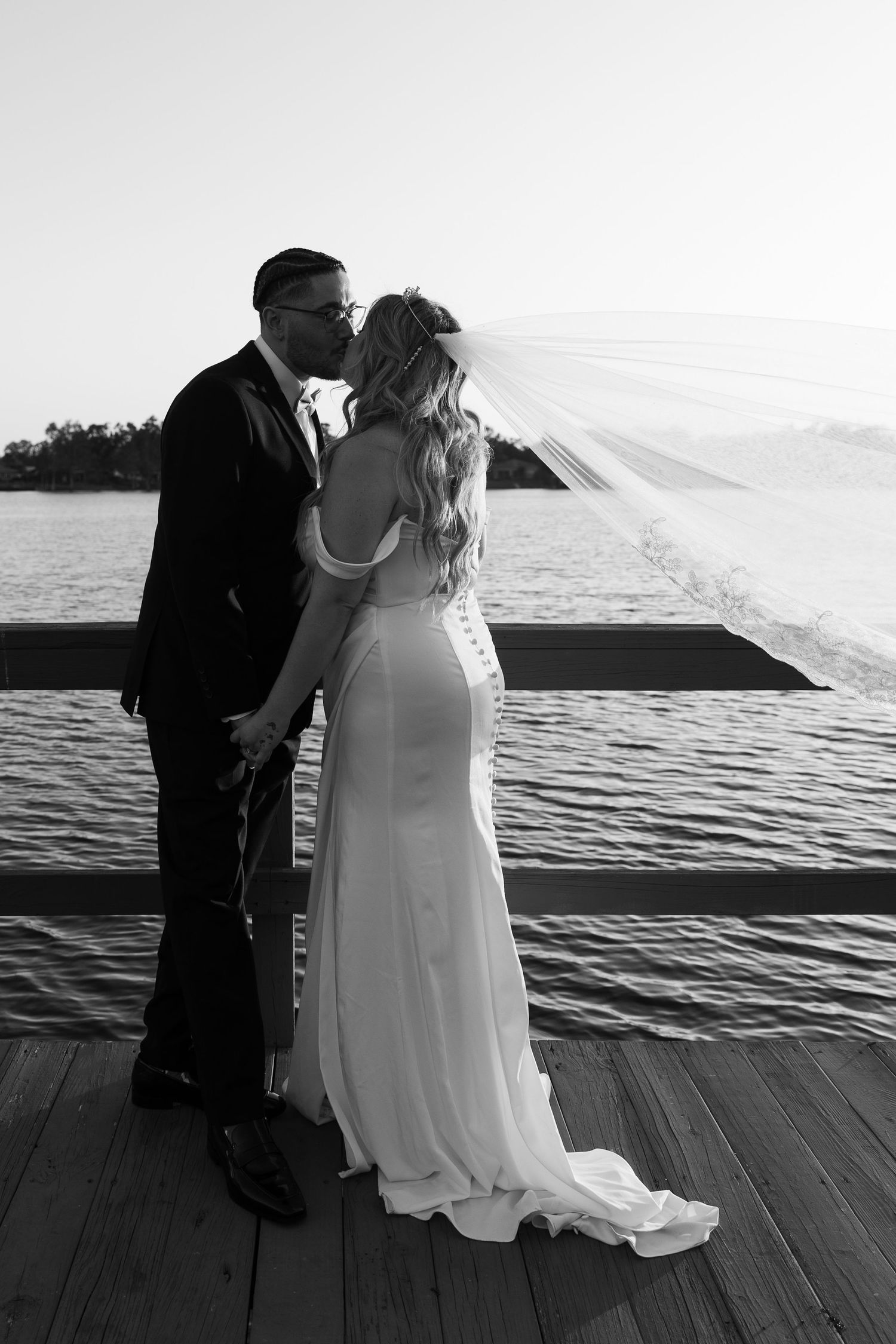 A black and white photo of a bride and groom kissing on a wooden dock by the water as her long veil blows in the wind.