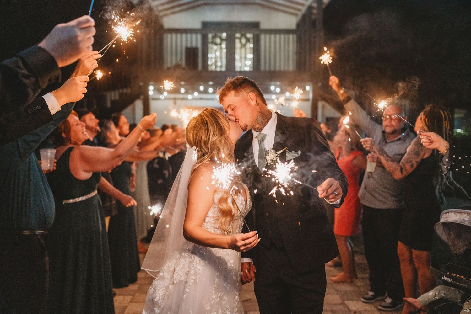 Newlyweds kiss while guests hold sparklers during an outdoor evening wedding exit.