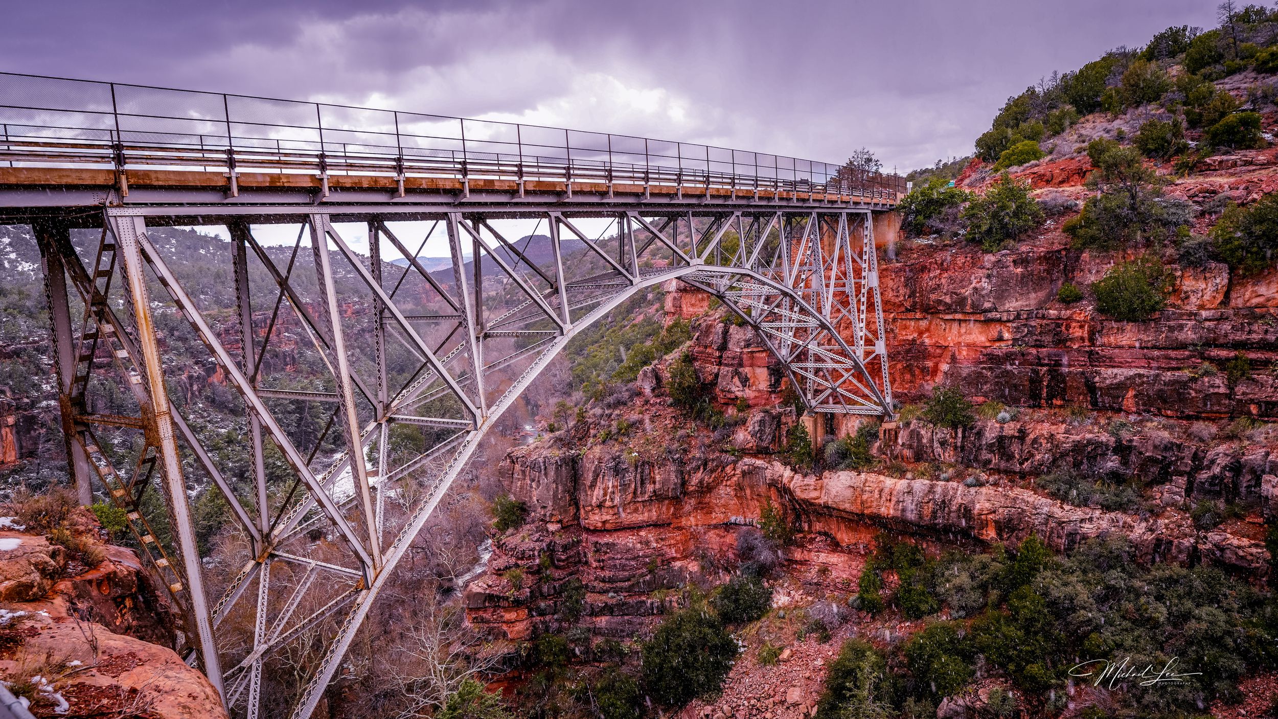 A steel arch bridge spans across a steep canyon with red rock walls under a dramatic purple sky.