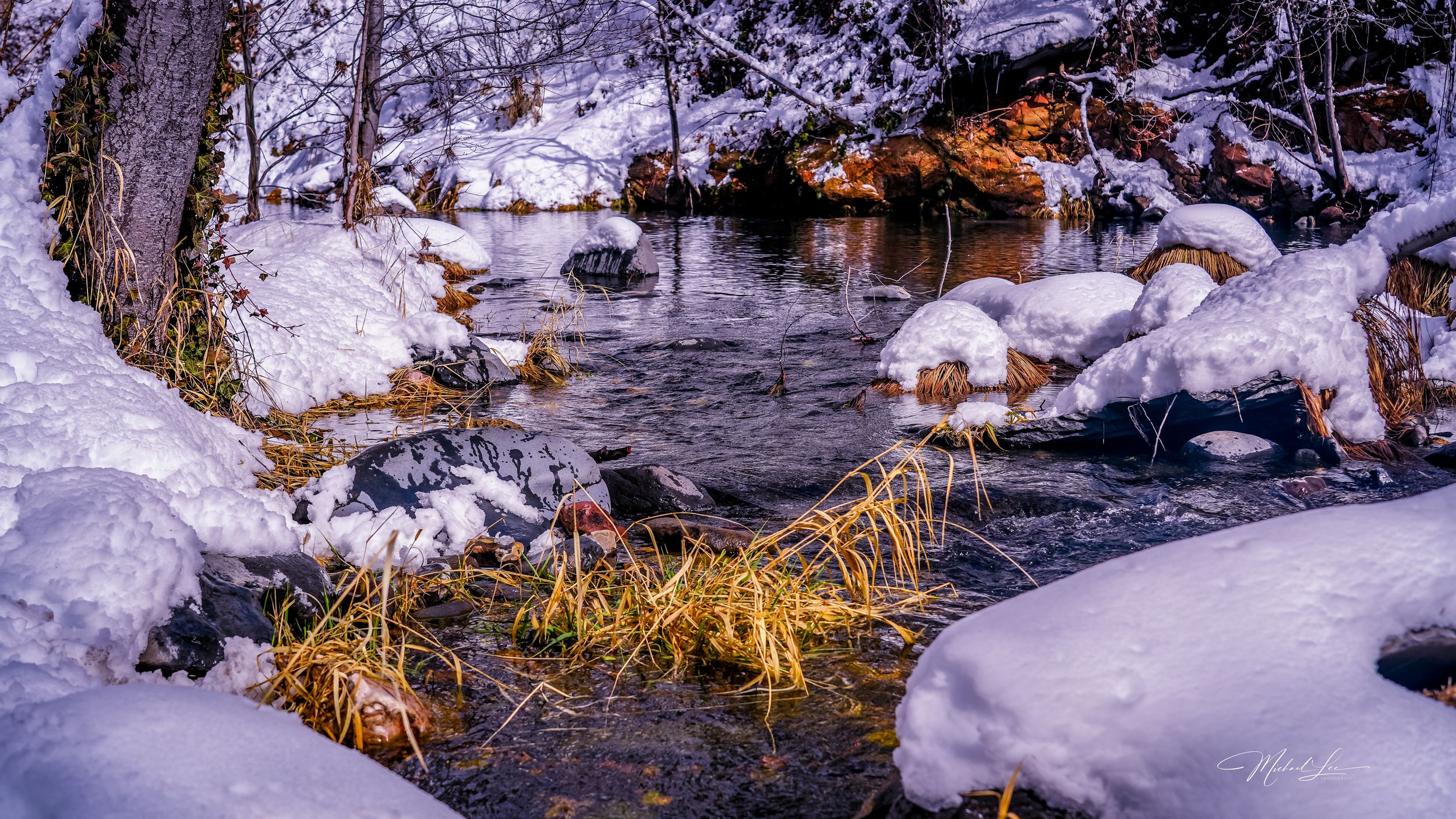 A flowing stream winds through snow-covered rocks and banks with golden grass peeking through the white landscape.