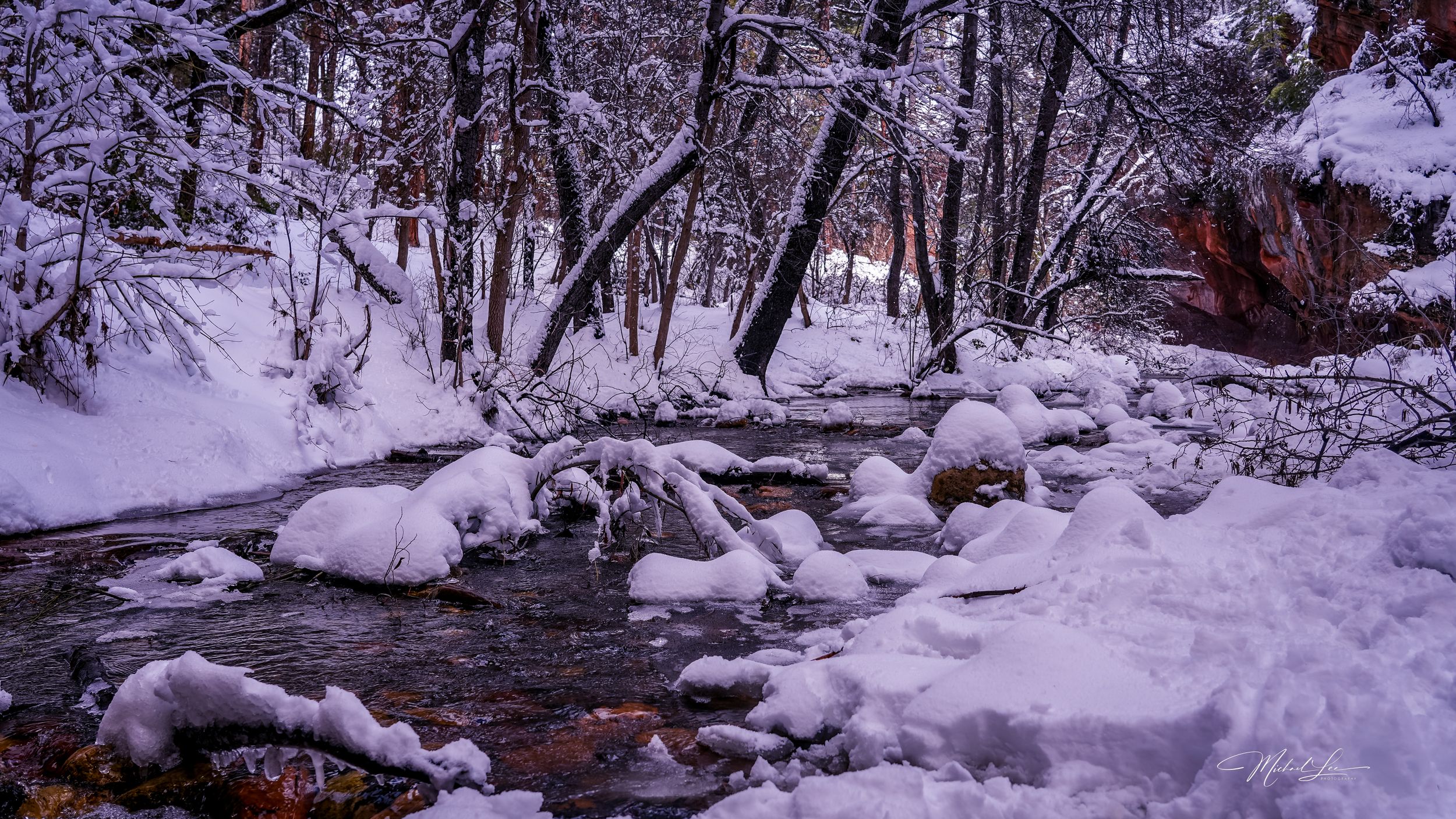 Snow-covered winter stream flows through a forest with rocks and bare trees creating a serene natural landscape.
