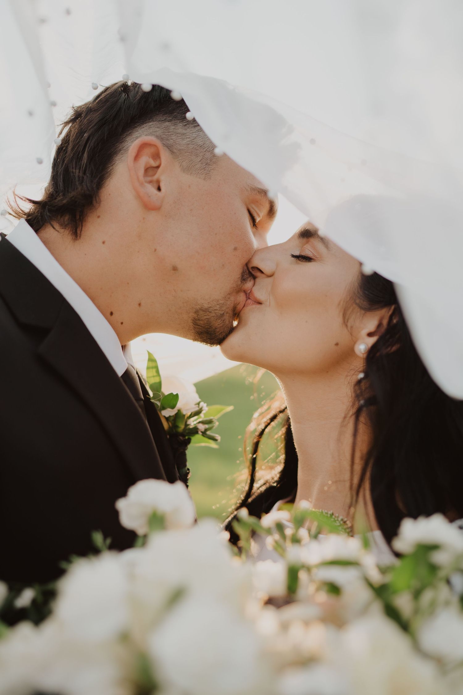 Romantic wedding kiss captured through white veil with flowers in foreground.