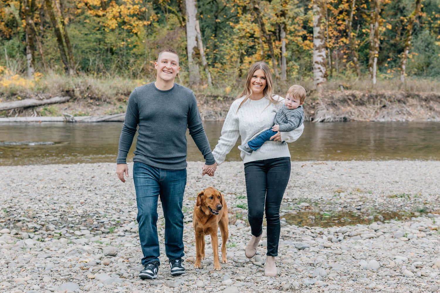 Happy family walking golden retriever dog along rocky riverbank in autumn setting.