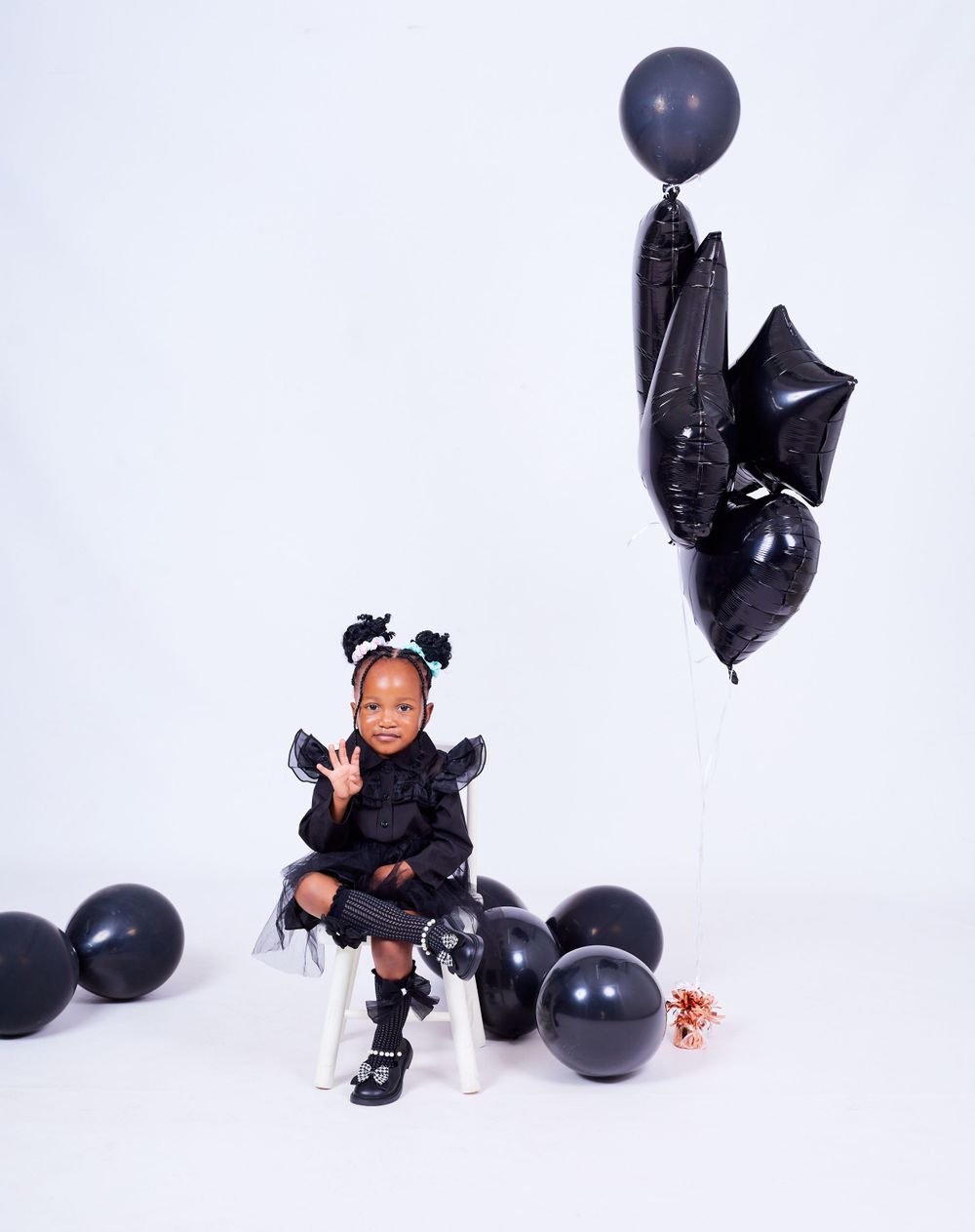 Child in black dress poses on white chair surrounded by elegant black balloons in studio setting.