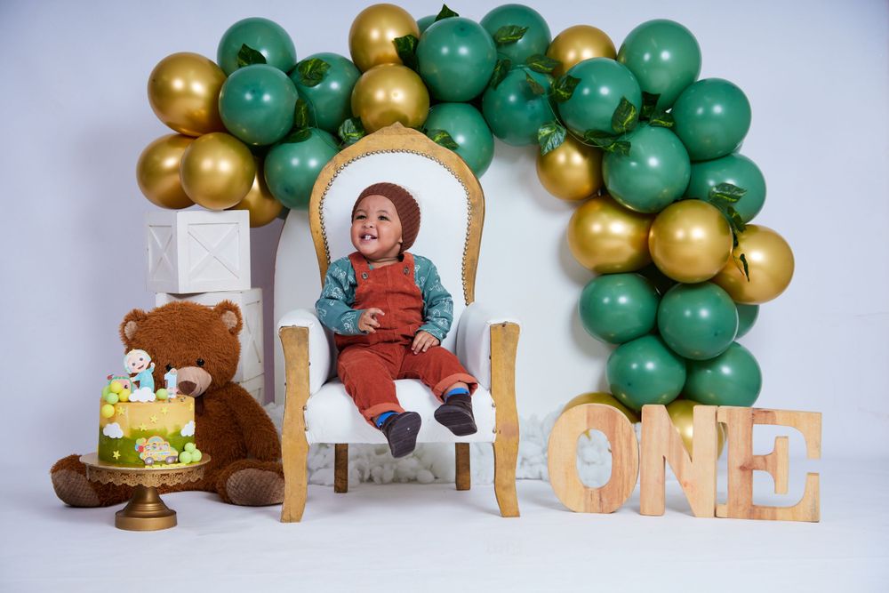 Birthday setup with gold and green balloon arch, white throne chair, teddy bear and cake display.