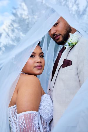 Elegant wedding couple poses against cloudy backdrop in white and burgundy attire.