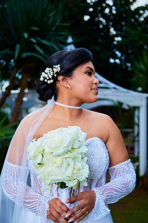Bride holds white rose bouquet in detailed beaded wedding gown outdoors.