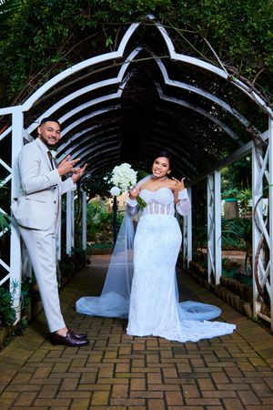 Newlyweds pose playfully under a white garden arbor with brick pathway.
