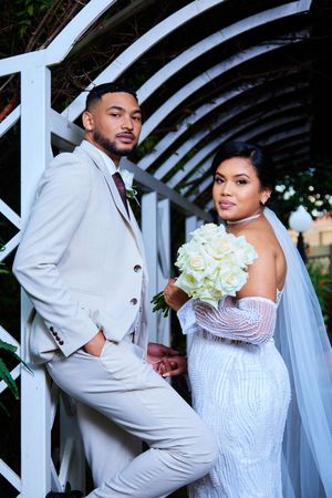 Wedding couple in white attire pose elegantly under an arched garden trellis.