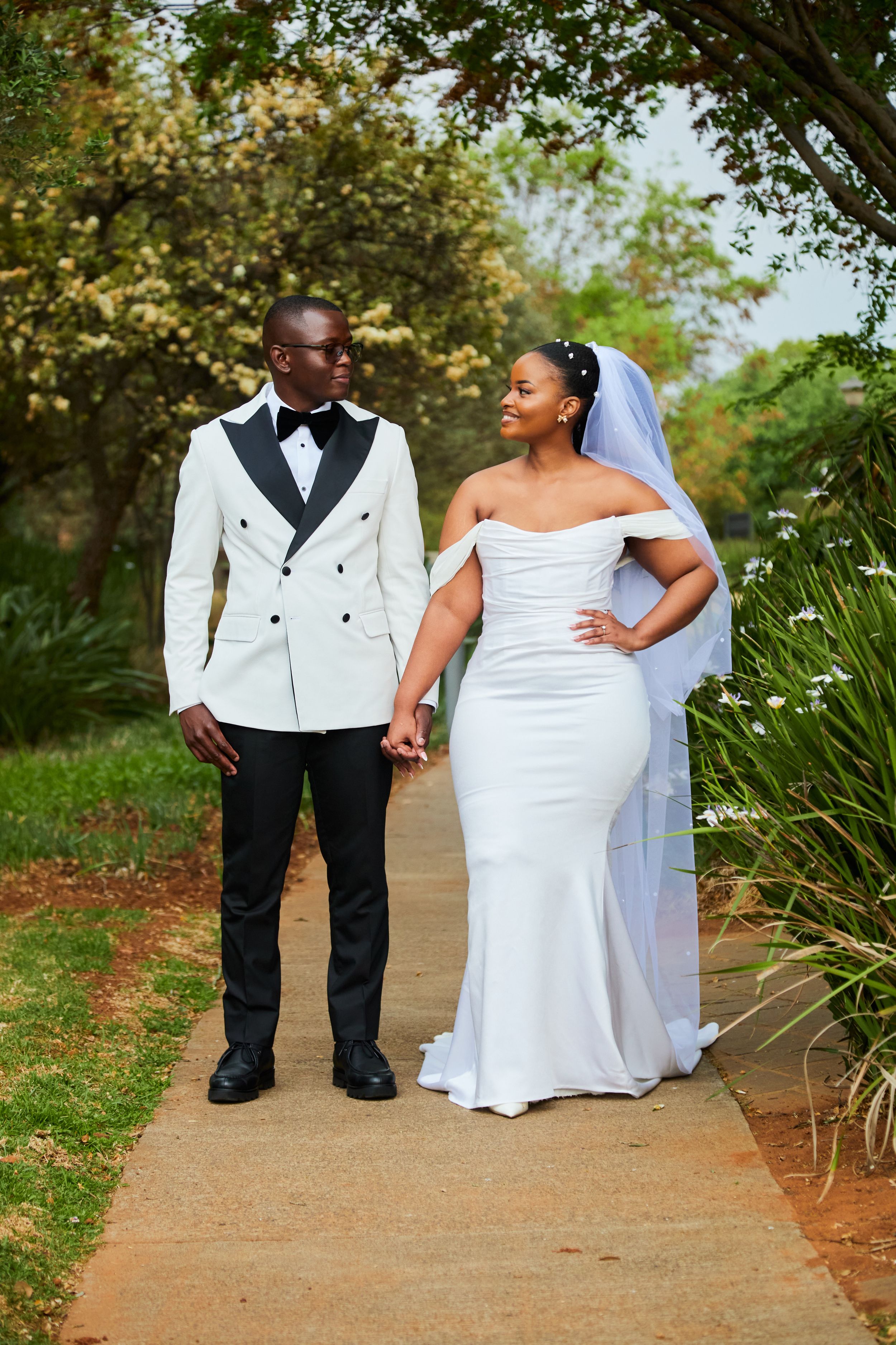 A couple in wedding attire walks together on a garden path surrounded by greenery.