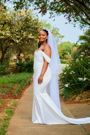 A bride in a flowing white wedding dress poses gracefully on a garden pathway with a long trailing veil.