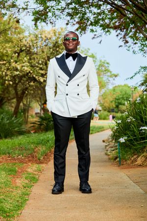 A groom in a classic white tuxedo and black bow tie poses on a garden pathway.
