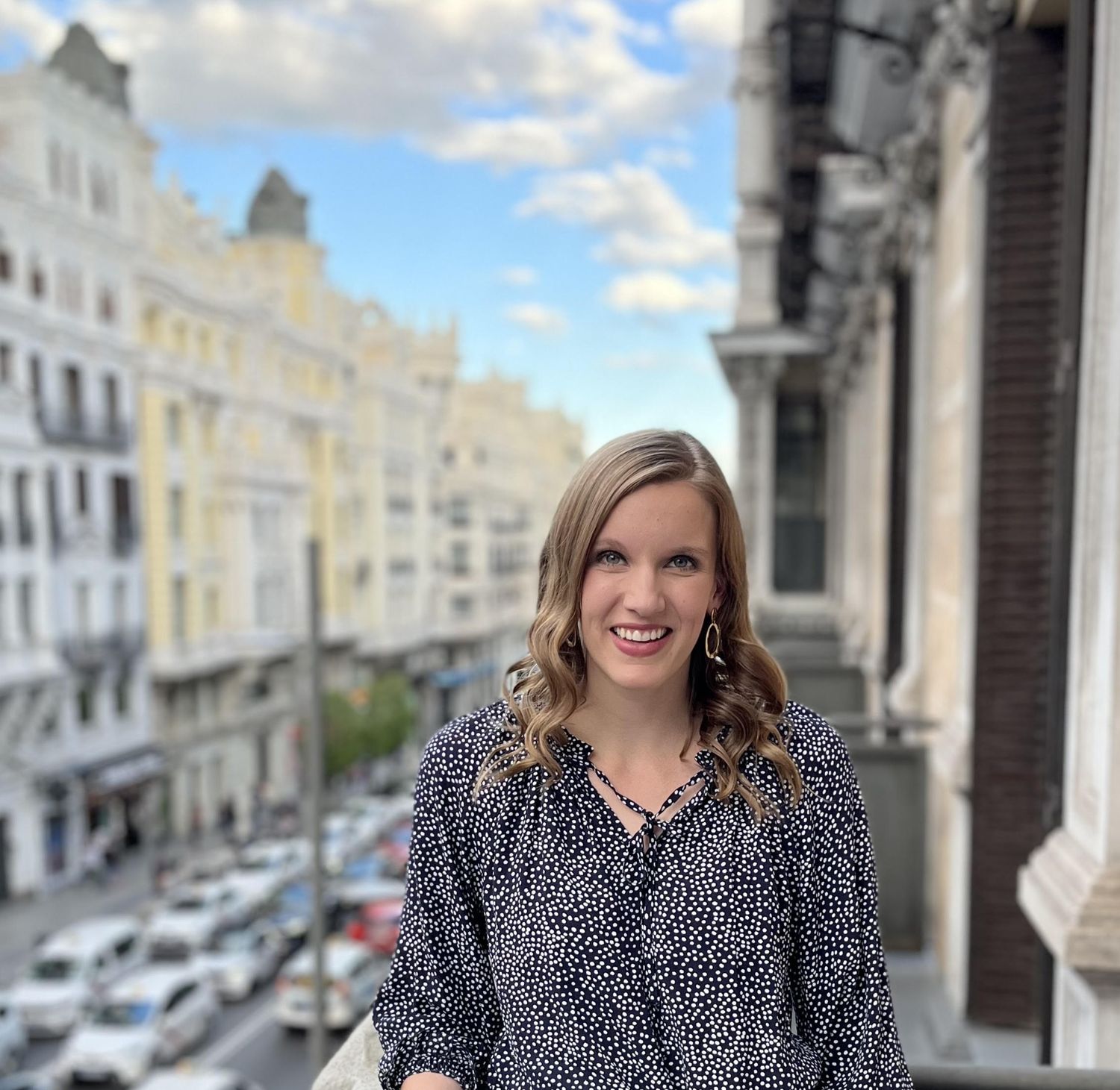 Woman in polka dot dress standing on balcony overlooking European city street.