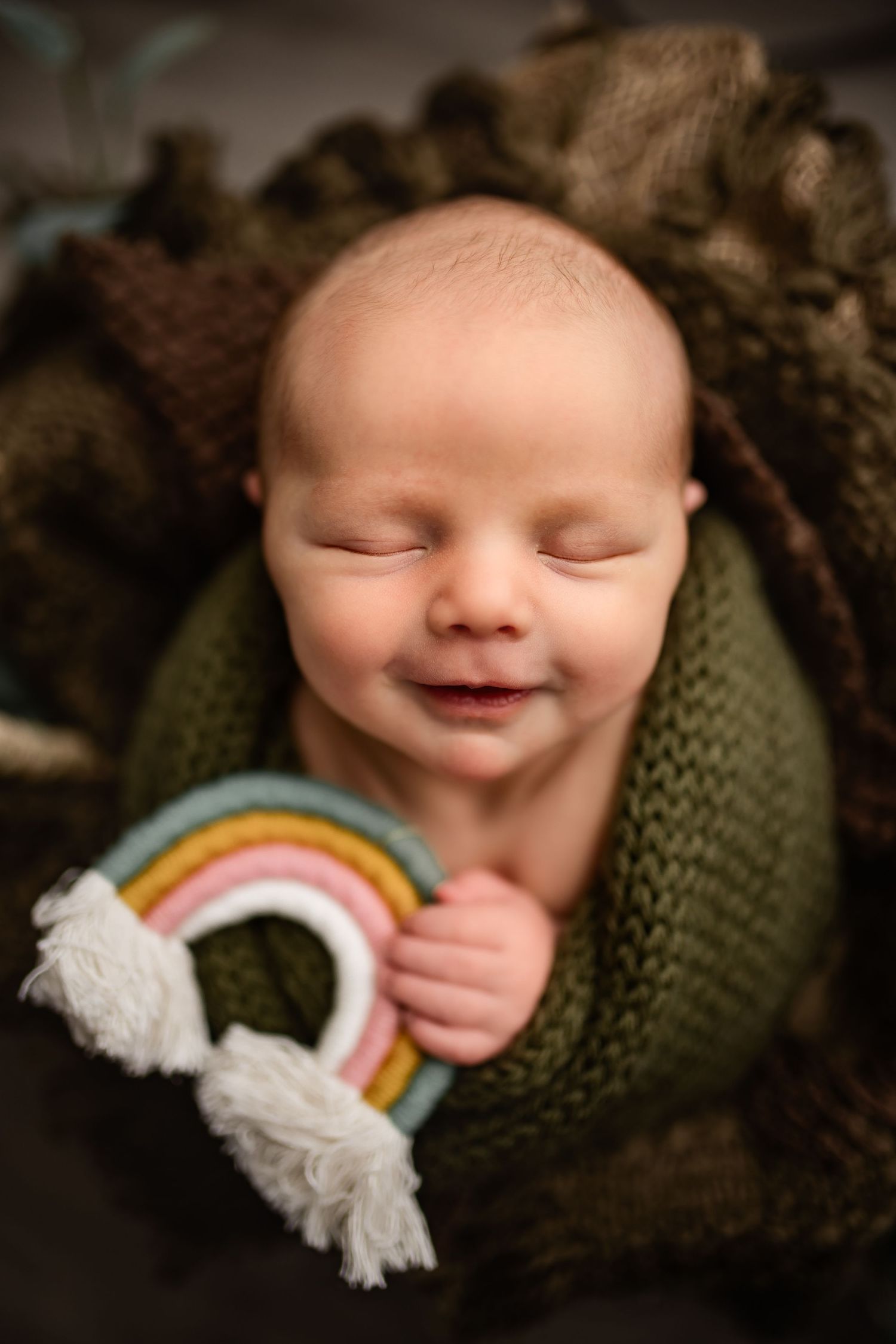 Smiling newborn baby boy holding a rainbow prop while swaddled in earth tones and posed in a newborn studio in Madison WI
