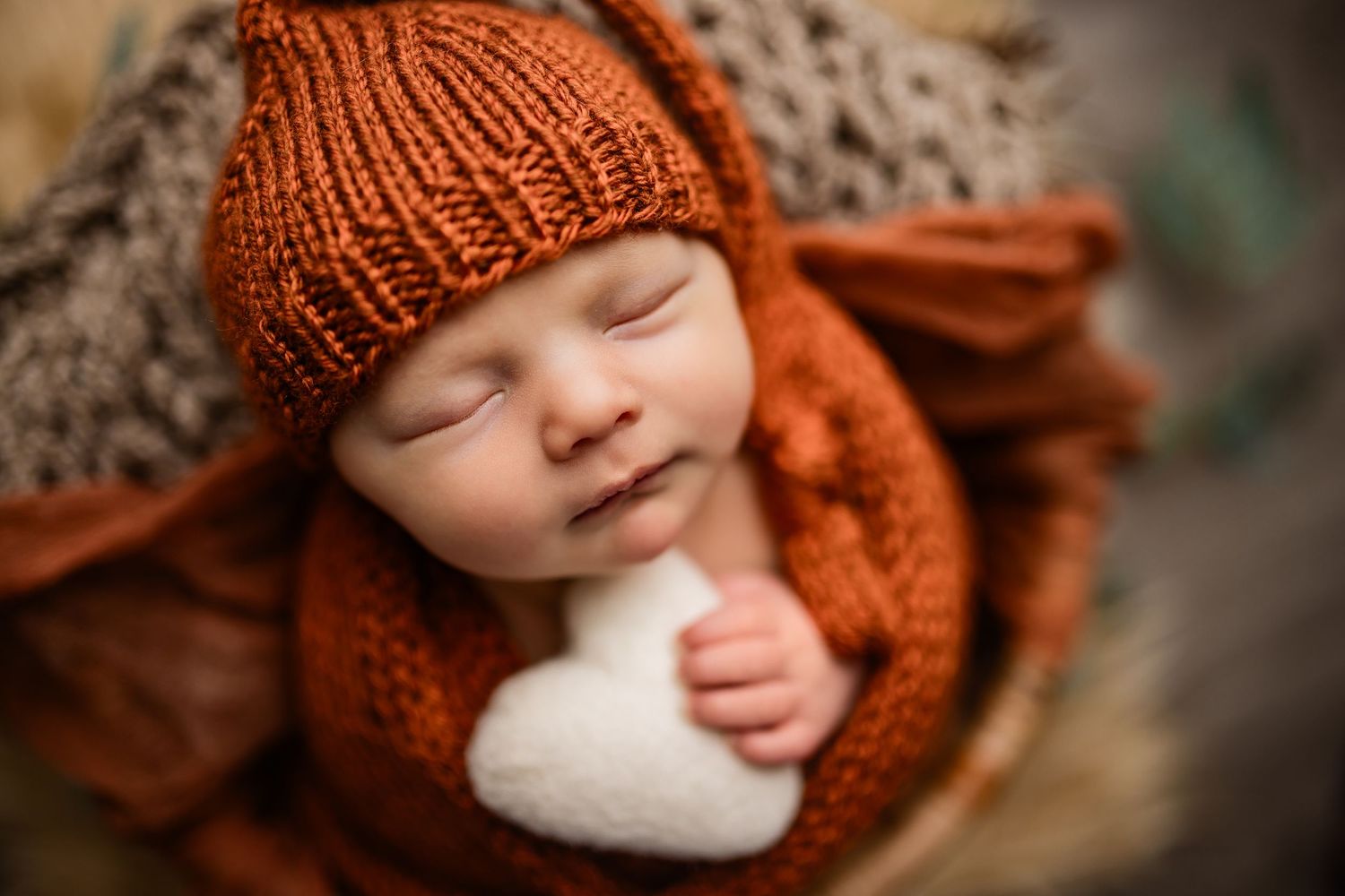 Sweet baby boy swaddled in an earth tone wrap with a sleepy cap holding a heart prop in a newborn studio in Waunakee WI