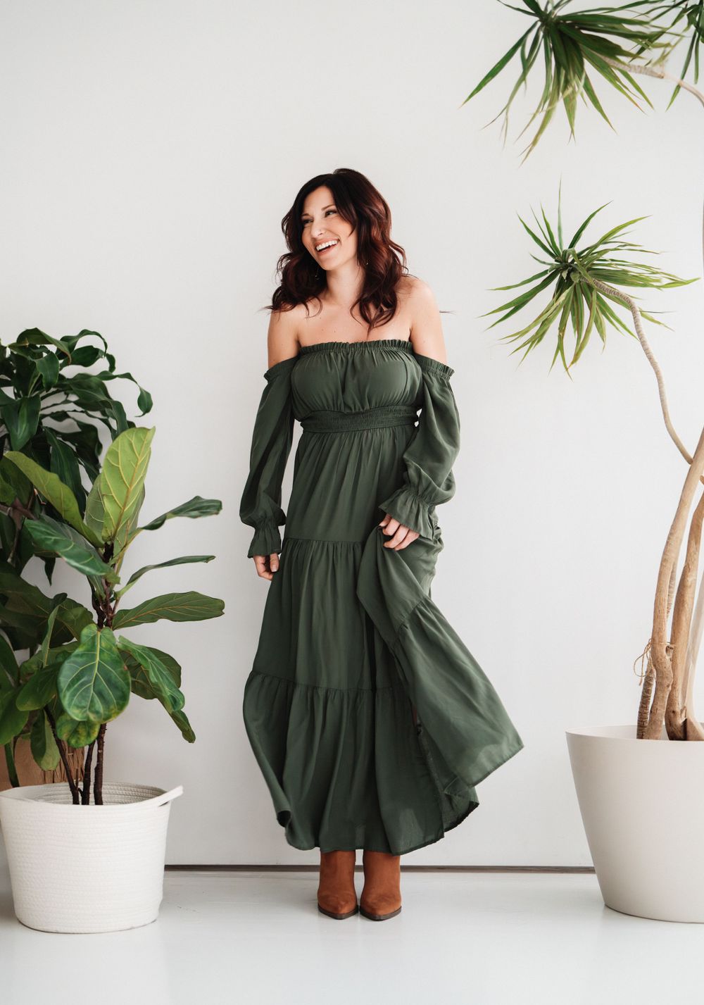 A smiling brunette woman poses in an olive green off-shoulder maxi dress surrounded by indoor plants in Cincinnati