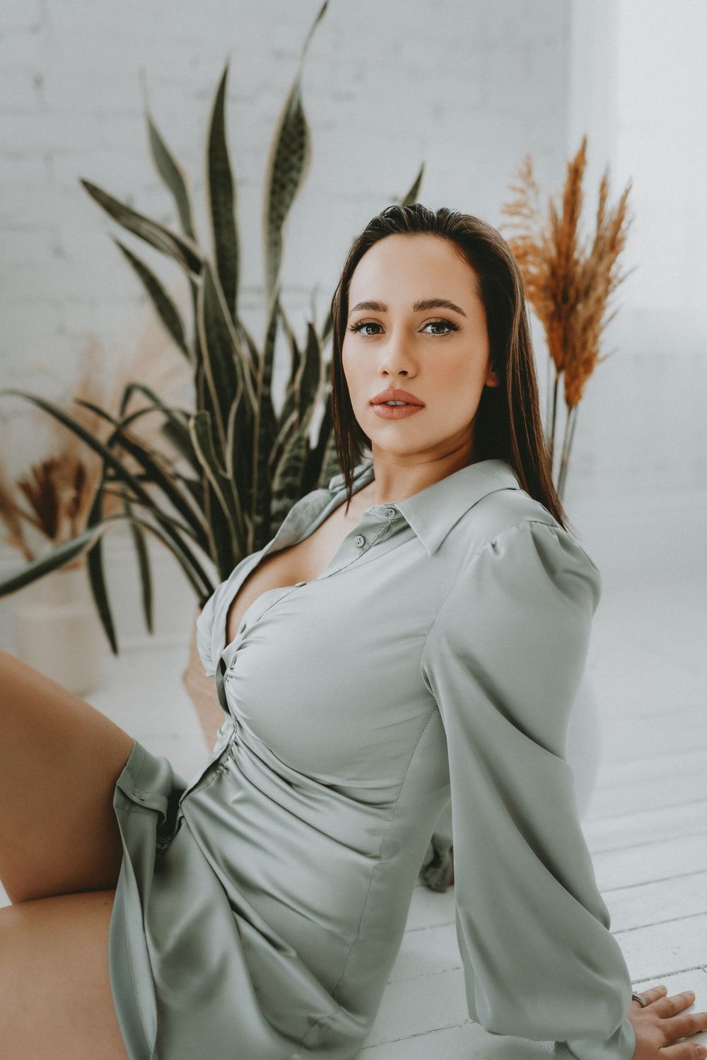Woman in sage green satin shirt dress poses gracefully against white brick wall with tropical plants in a Cincinnati studio