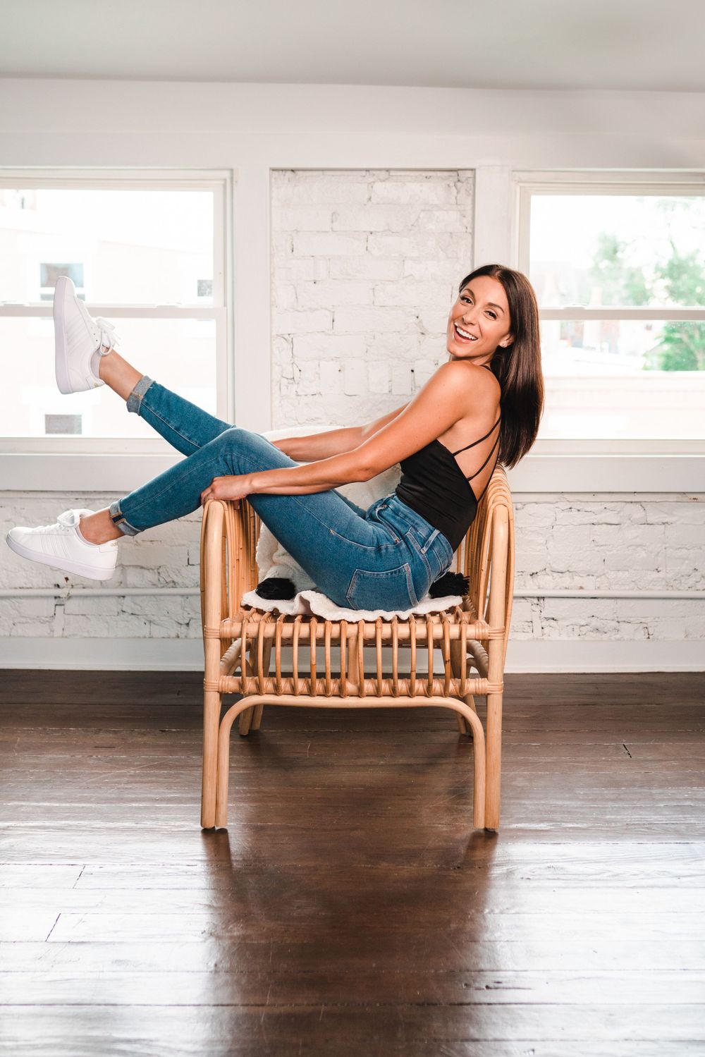 A smiling brunette woman sits casually in a rattan chair wearing jeans and a black top in a bright room in Cincinnati