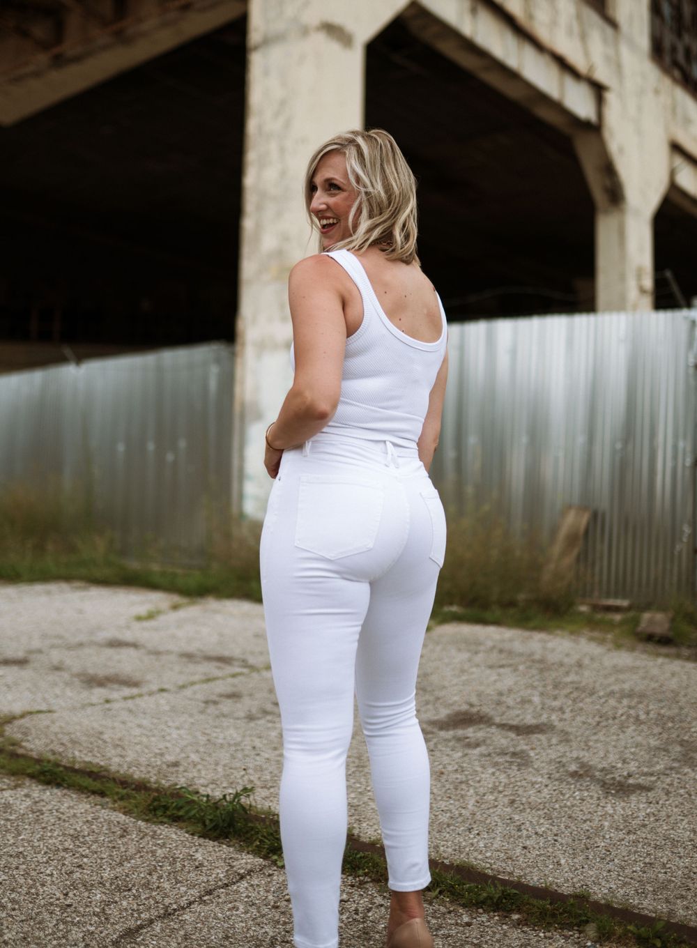 A blonde woman poses outdoors wearing a white tank top and fitted white jeans near an industrial building in Cincinnati