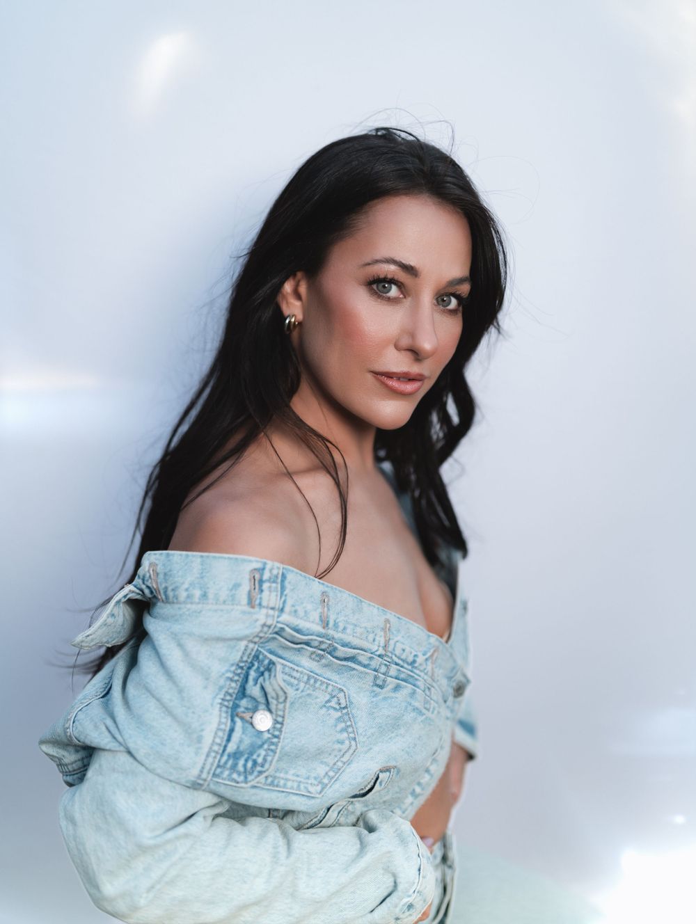 Brunette woman in off-shoulder denim jacket posing against white background with dark flowing hair in Cincinnati