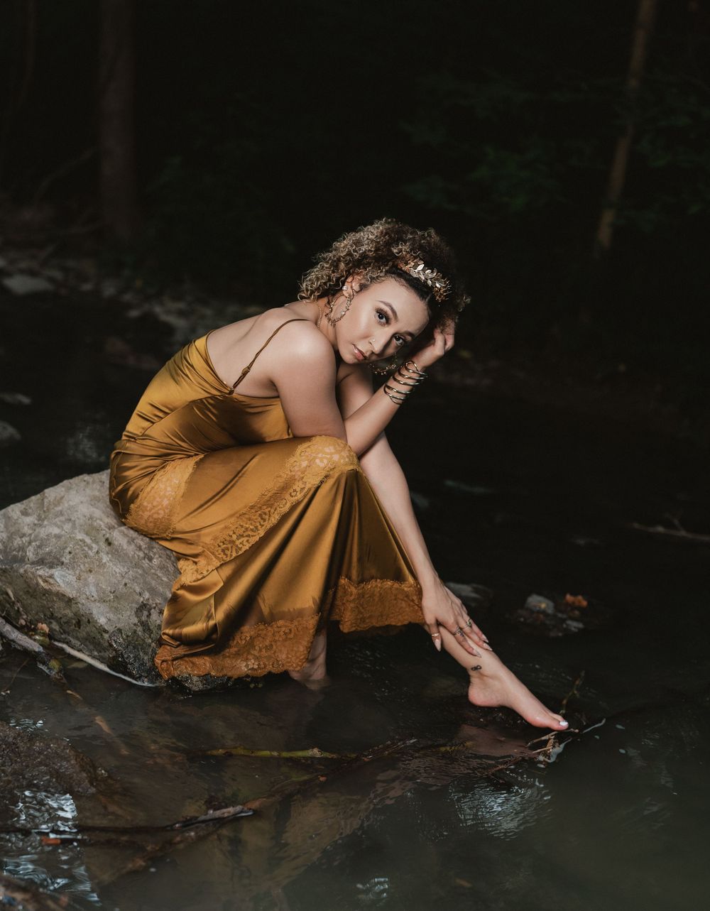 Young woman in a tiara and golden dress sitting on a boulder inside a creek in Cincinnati Ohio for her birthday photoshoot