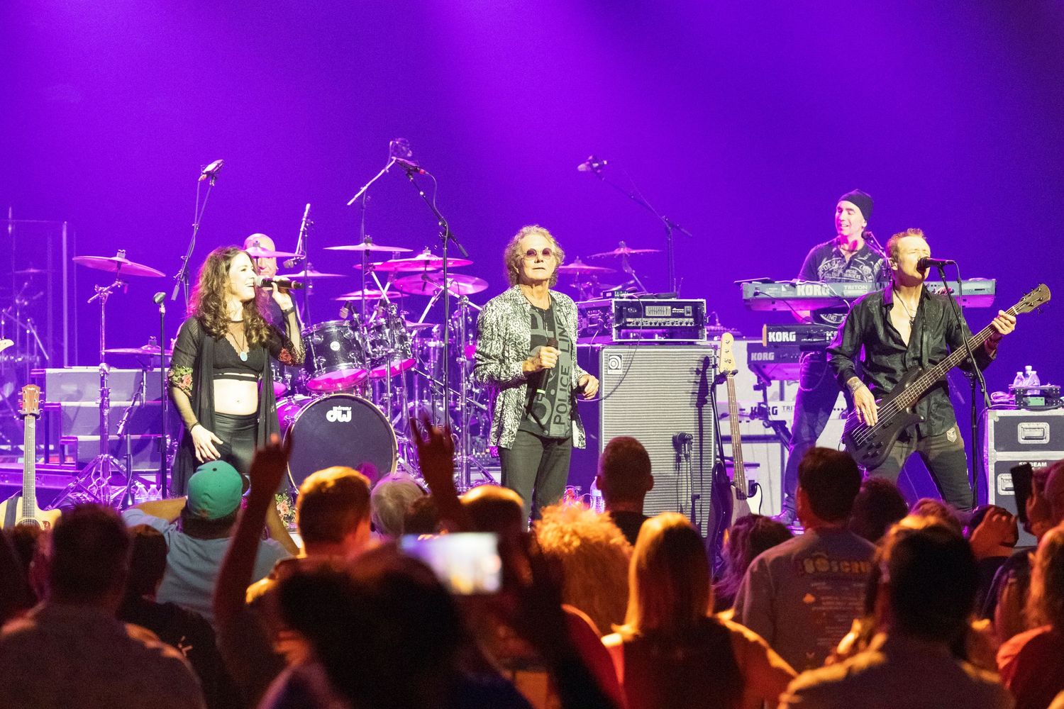 Rock band performing on stage with purple lighting and energetic crowd in the foreground at a live concert.